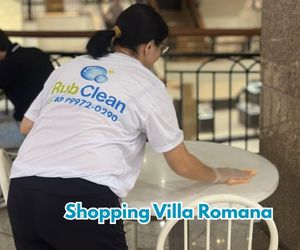 Funcionária da Rub Clean com camiseta branca — logo e telefone visíveis — higieniza uma mesa redonda em área interna do Shopping Villa Romana.