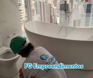 Técnico da Rub Clean usando capacete verde limpa o banheiro de um apartamento de alto padrão da FG Empreendimentos; banheira moderna e vista para prédios ao fundo.